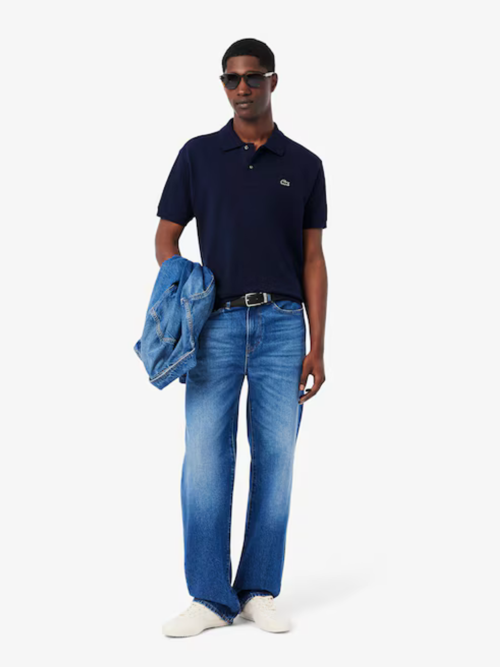 Man wearing a navy polo shirt and blue jeans holding a denim jacket on a white background